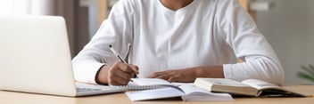 A young woman is sitting at a table with a laptop computer and several books and notebooks, writing in a notebook with a pen.