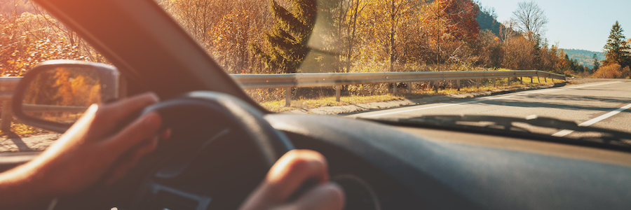 Person's hands on steering wheel of car, driving down a highway with mountains and trees in the background